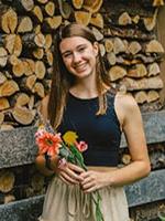 Girl with brown haor holding flowers standing in front of stacked wood